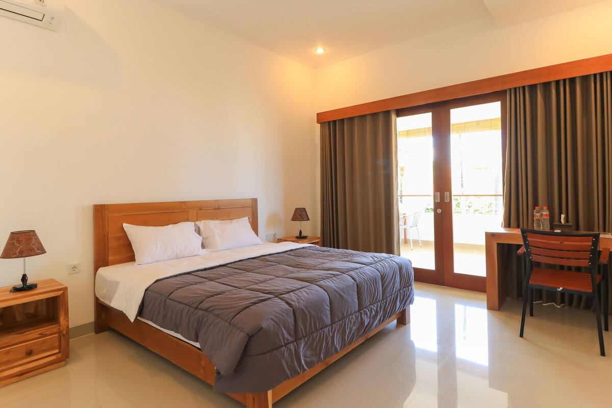 A hotel room with a wooden bed, a desk with a chair, and a view of the outdoors through a sliding door.