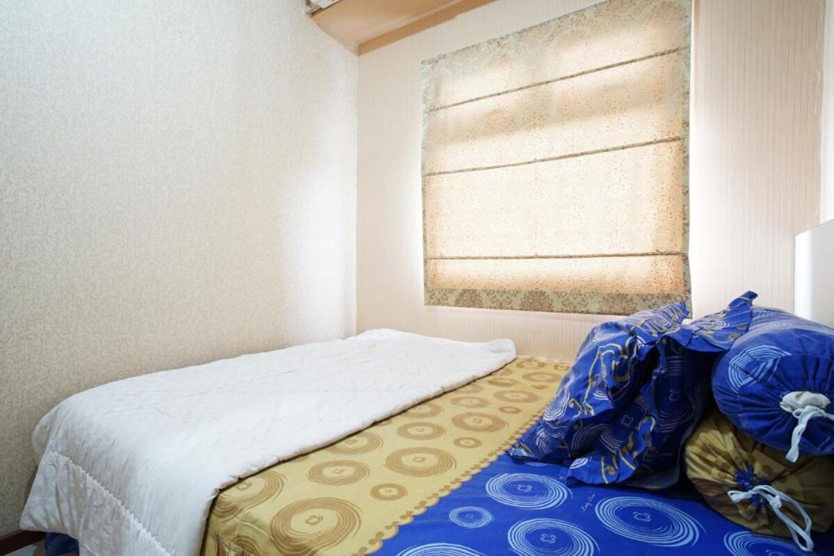 A single bed with a white blanket and patterned bedding, a window with blinds, and a patterned wall.
