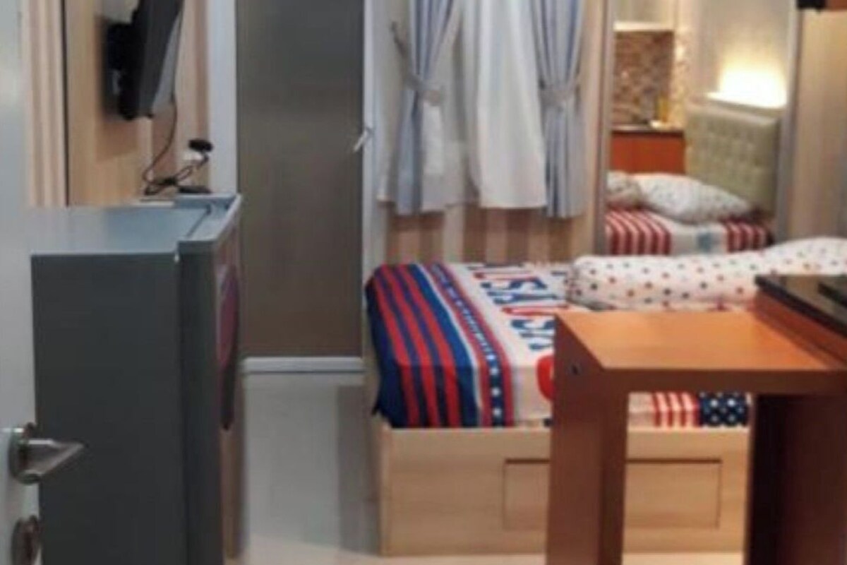 A compact hotel room with a kitchenette, a bed with a striped blanket, a small table, and a desk.
