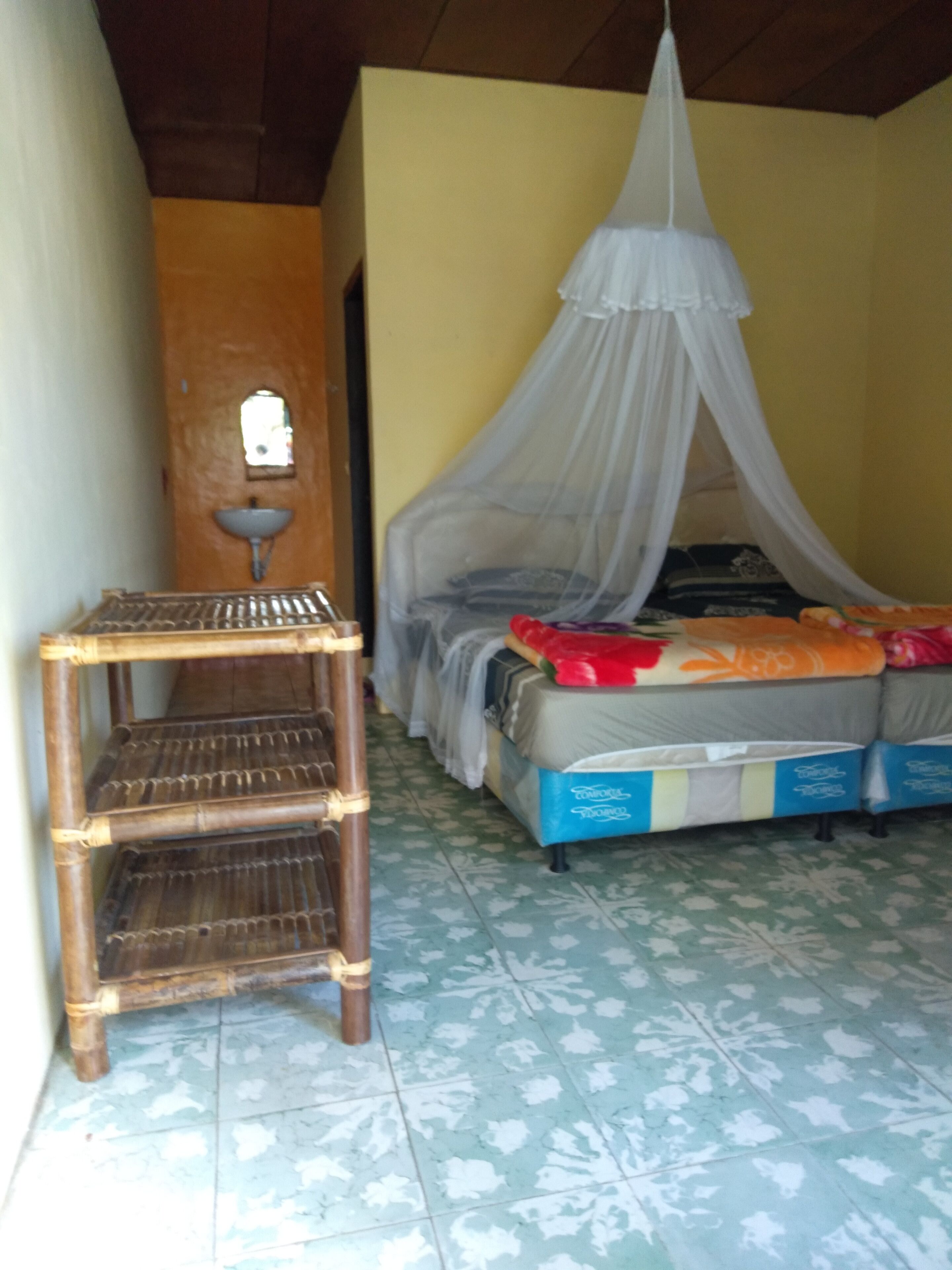 A room with a bed under a mosquito net, a small shelf, and a sink on the wall.