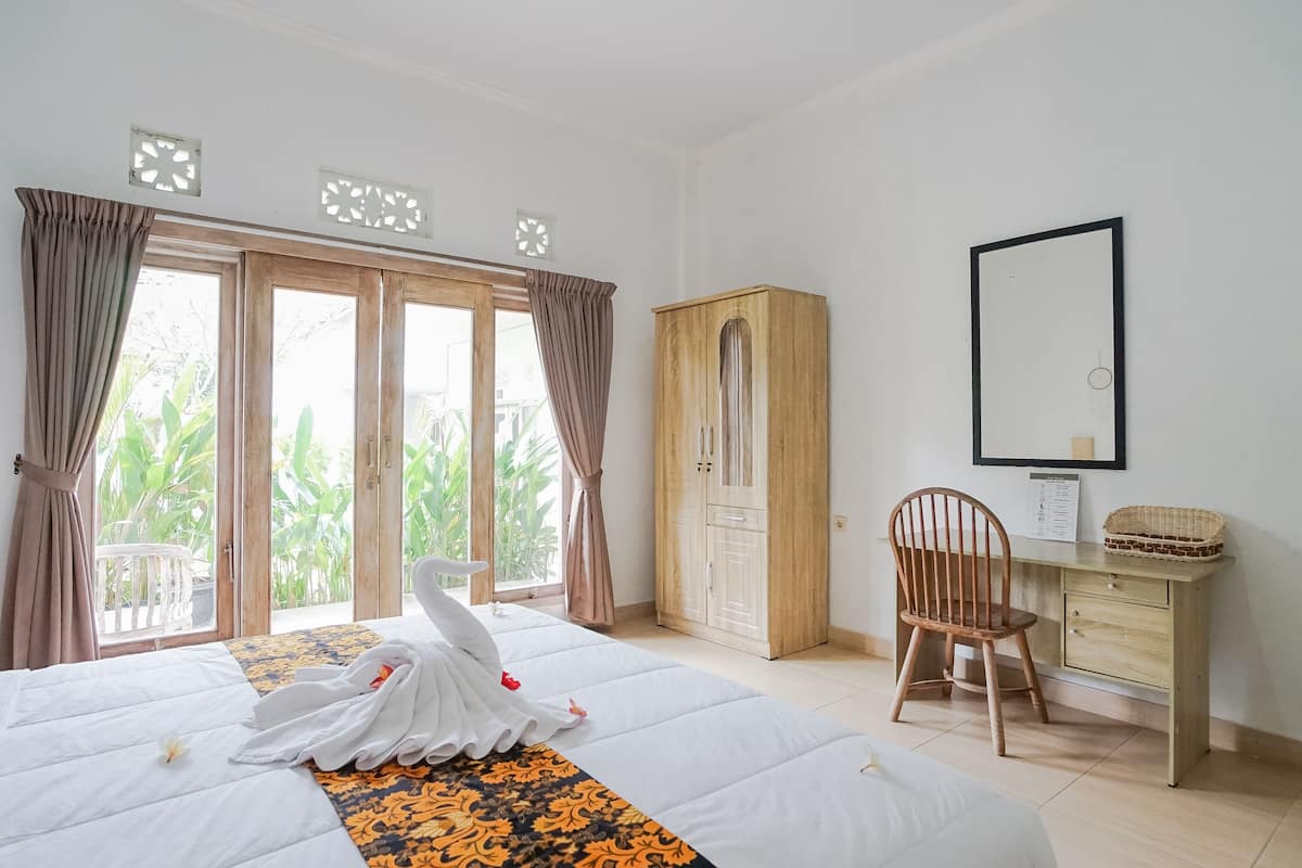 Quarto de hotel com cama, guarda-roupa de madeira, secretária e uma cadeira, e uma janela grande com cortinas.