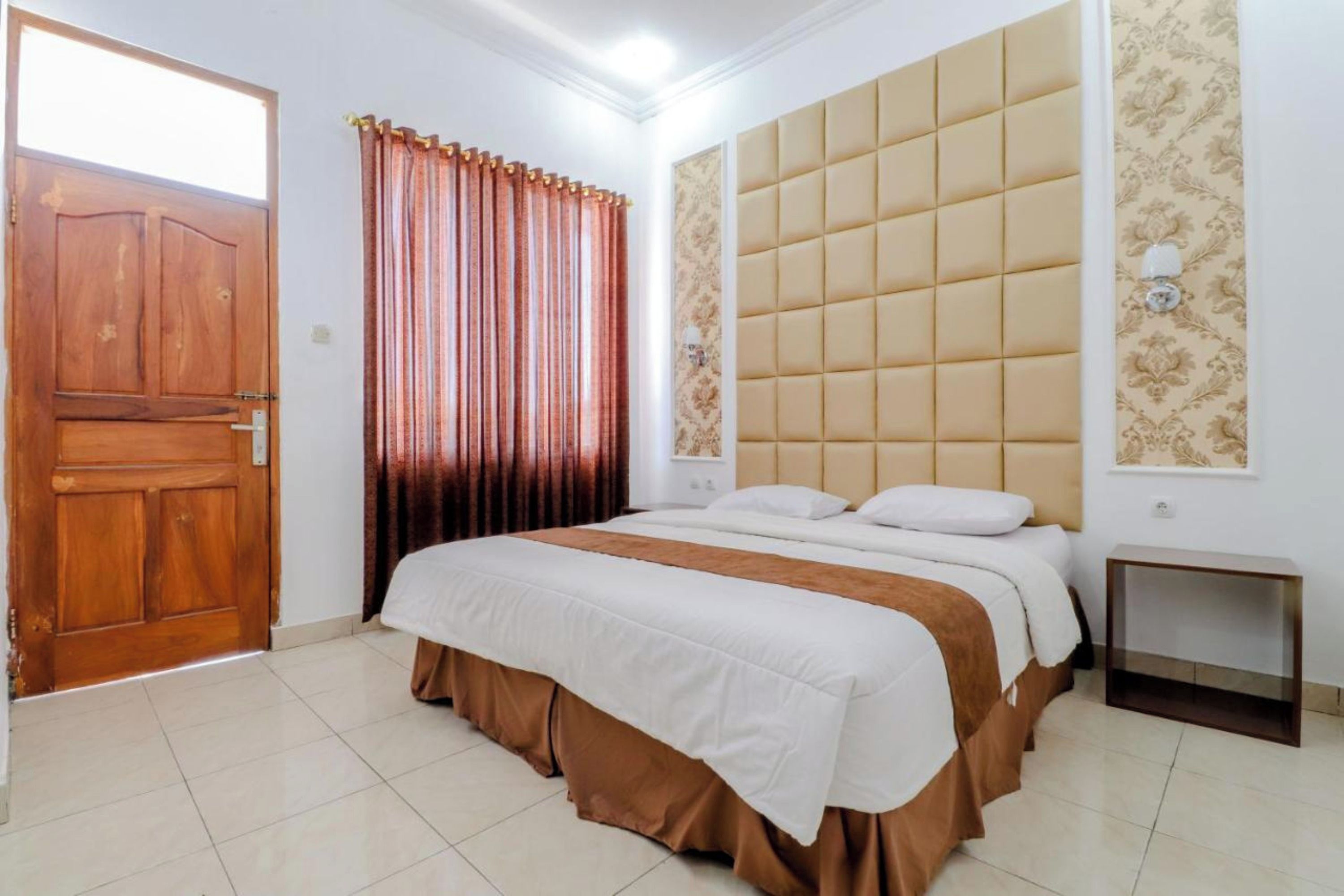A bedroom with a wooden door, a bed with a white and brown bedspread, a headboard with a tufted design, and a small bedside table.