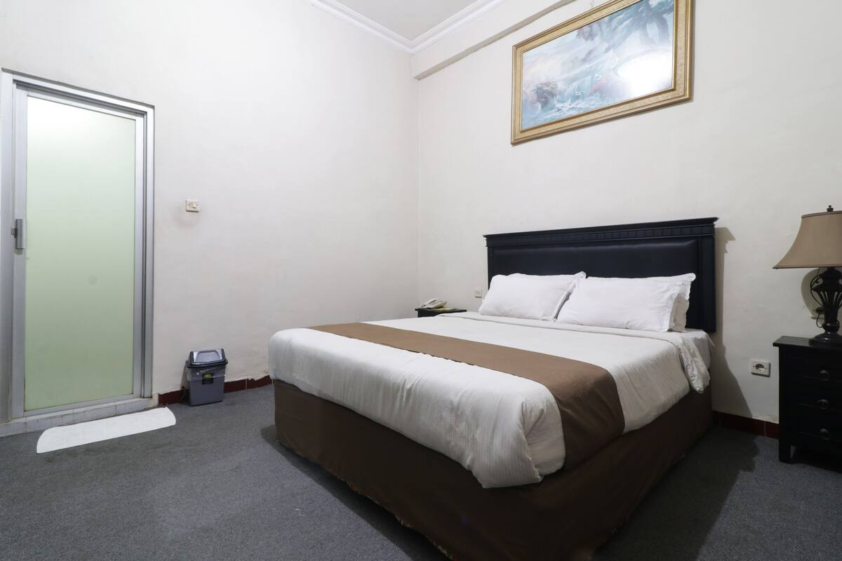 Una habitación de hotel con una cama, una mesita de noche, una lámpara, un cubo de basura y una puerta.