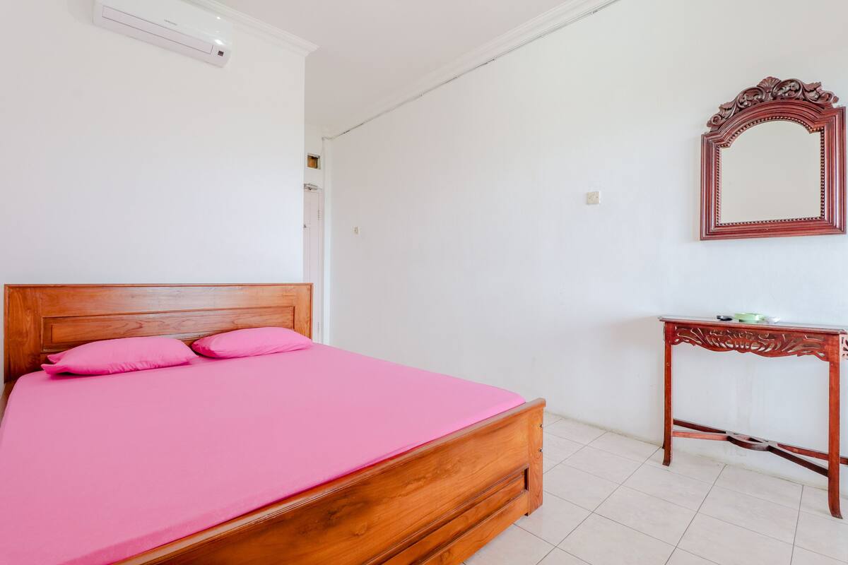 Un dormitorio con una cama de madera, ropa de cama rosa, una mesita de noche de madera y un espejo en la pared.