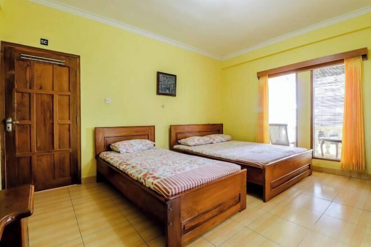 Una habitación con dos camas de madera, una puerta de madera, una ventana con cortinas y una pequeña mesa.