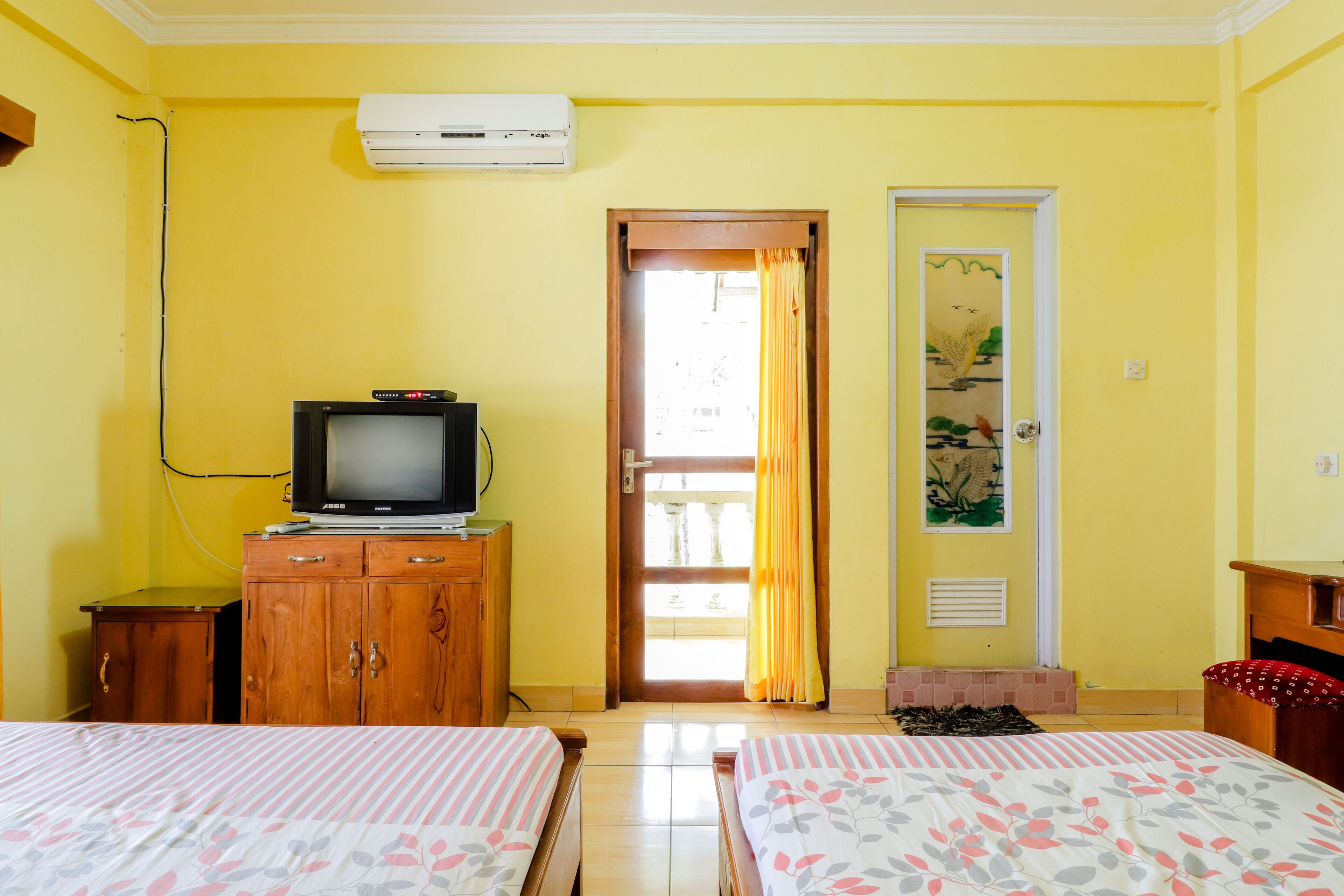 A room with two beds, a television on a wooden cabinet, an air conditioner on the wall, and a door leading outside.