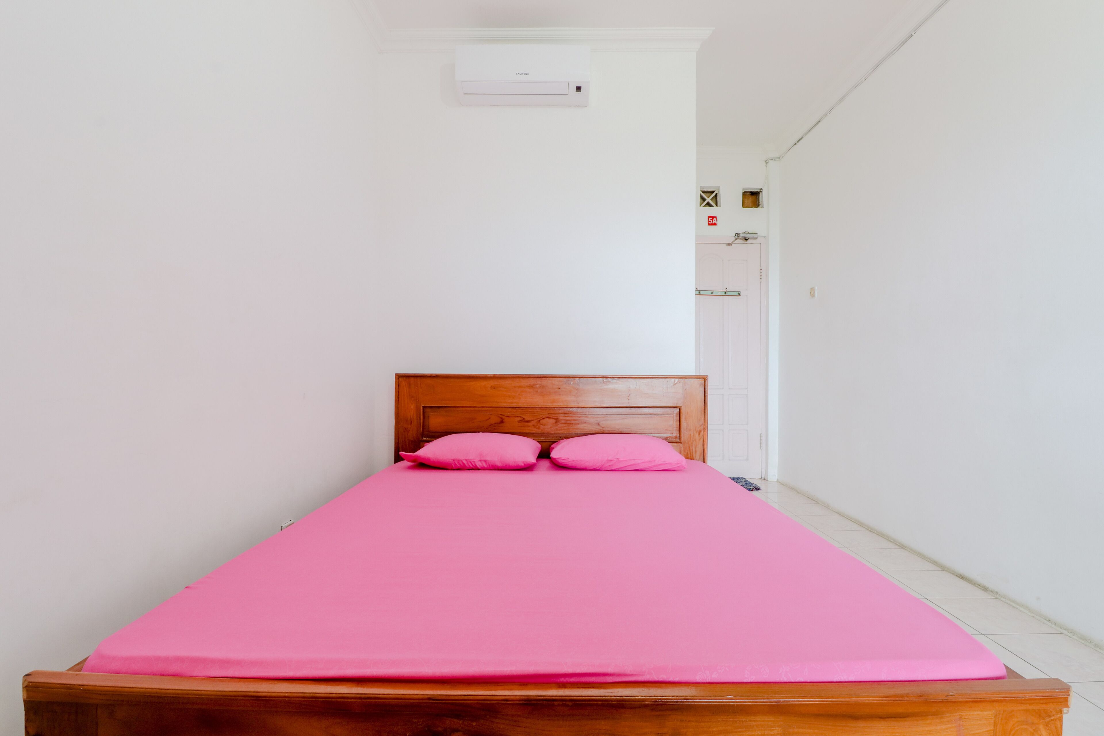 A single bed with a wooden headboard and pink bedding in a room with white walls and a tiled floor.