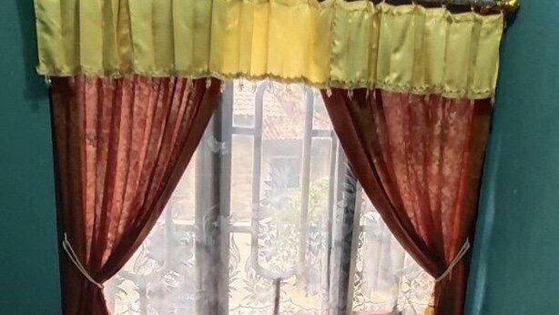 A bed with a red mattress and pillows, a window with yellow and brown curtains, and a view of a balcony with a railing. - Mas Bangkrenx Homestay (Depok)