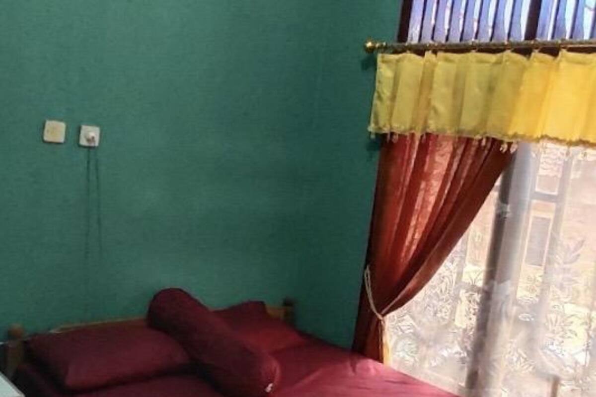 A single bed with a red bedspread and two pillows, a nightstand with a lamp, and a window with a yellow curtain.