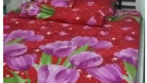 Uma cama com colcha floral vermelha e roxa, cabeceira de metal branca e parede branca ao fundo.