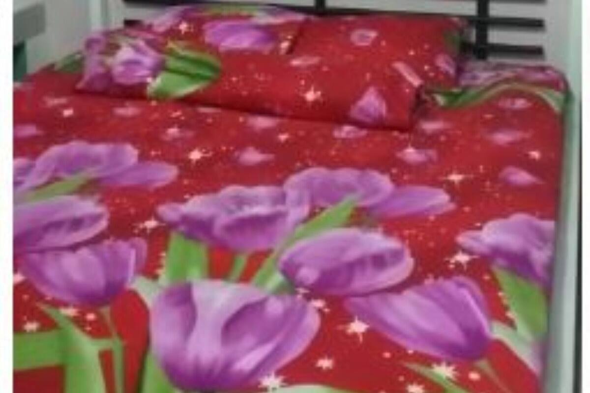Una cama con una colcha floral roja y morada, un cabecero de metal blanco y una pared blanca al fondo.