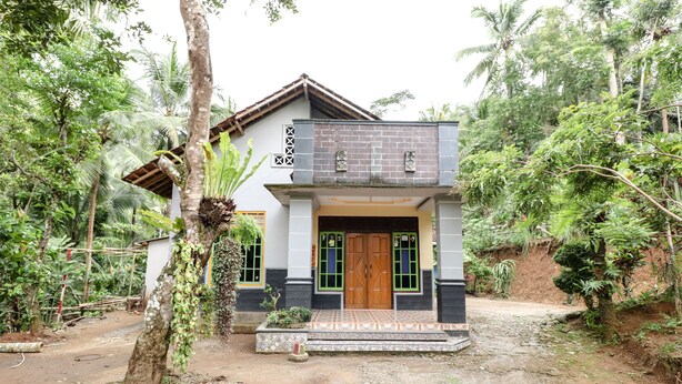 A two-story house with a tiled entrance and a wooden door, surrounded by lush greenery.