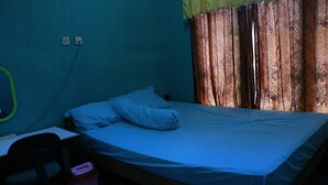 A single bed with a blue mattress and pillows, a desk with a chair, a trash bin, and a window with patterned curtains. - Sarjuni Homestay - Hostel (Kokap)