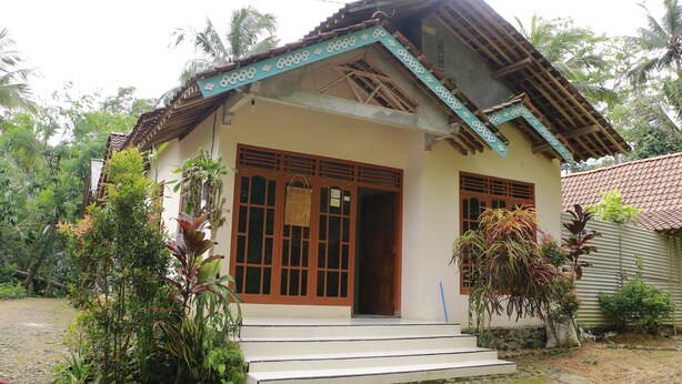 A two-story house with a tiled roof, a front porch, and a small garden area with various plants. - Suparman Homestay - Hostel (Kokap)