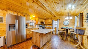 A wooden cabin interior with a kitchen featuring a stainless steel refrigerator, wooden cabinets, a central island with stools, and a dining table. - Broken Bow Abode w/ Hot Tub < 7 Mi to Lake! (Broken Bow)