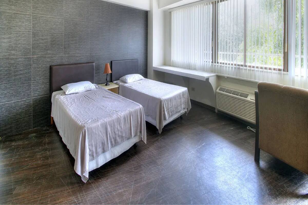 Habitación de hotel con dos camas, un escritorio, una silla y un ventanal con persianas.