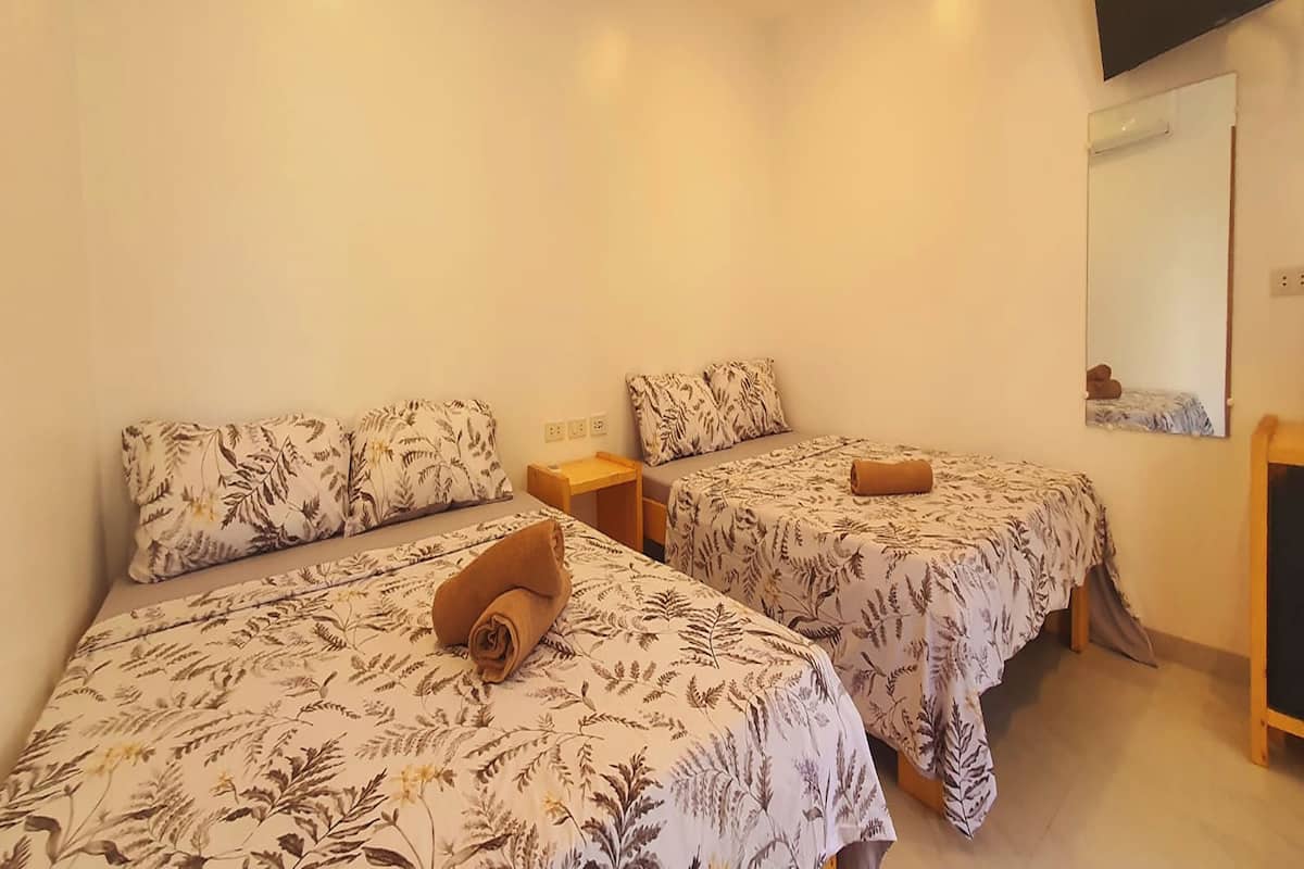 A hotel room with two beds, each with a floral bedspread and a rolled towel on one bed. There is a small wooden nightstand between the beds, and a mirror on the wall.