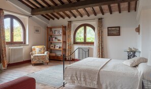 A cozy bedroom with a bed, a chair, a bookshelf, and two windows.