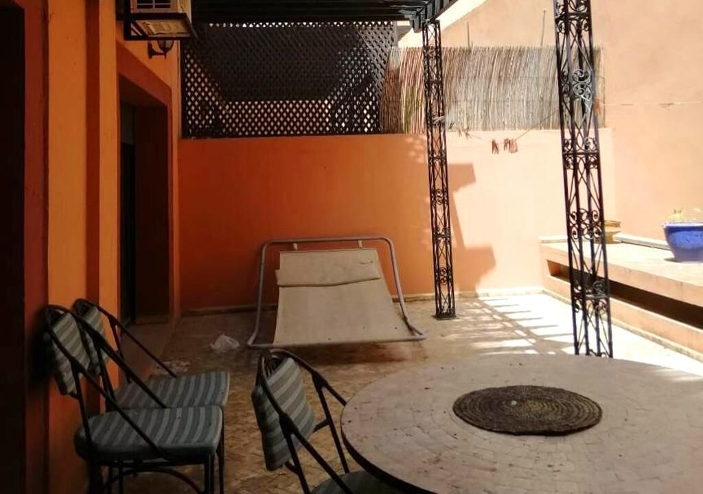 Een zonnig terras met een ronde tafel, twee stoelen, een kleine hellingbaan en een decoratieve metalen balustrade.