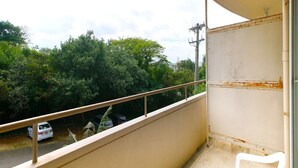 Un balcon avec une chaise en plastique blanche, une rambarde en métal et une vue sur une route bordée de voitures garées et d’arbres.