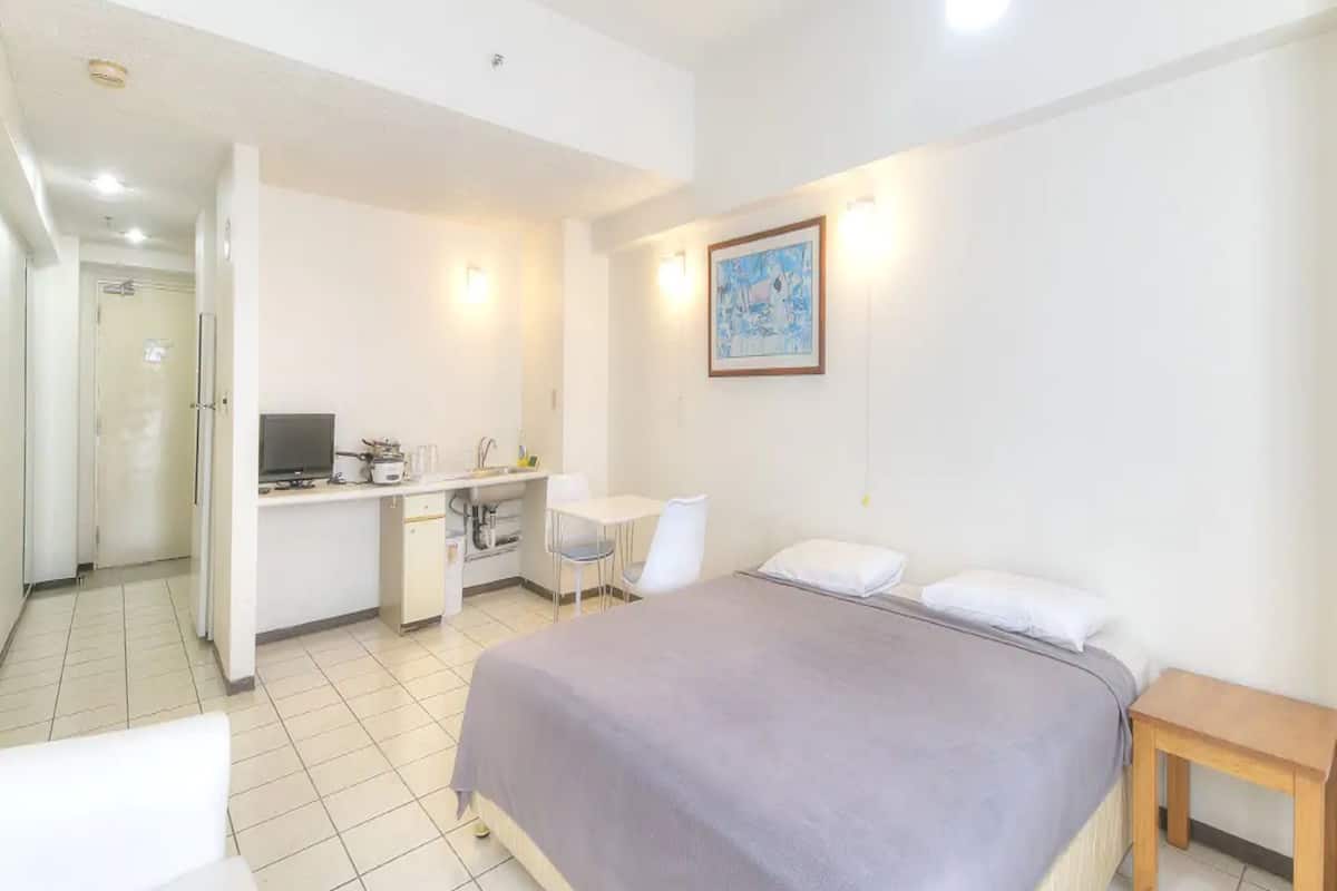 A compact hotel room with a bed, a small desk, a chair, and a kitchenette.