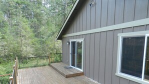 A wooden deck with a glass door leading to a covered area, surrounded by a forested landscape. - Anderson Island's most peaceful home (Anderson Island)