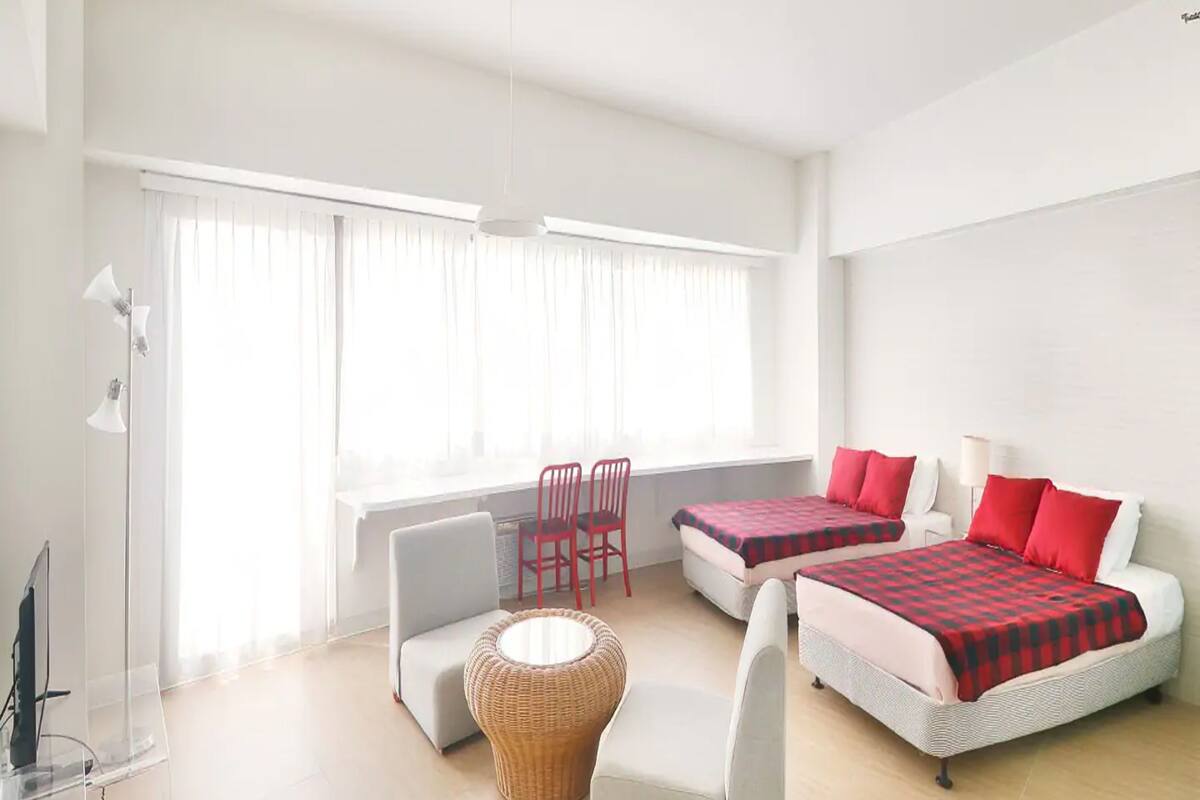 A bright, modern room with two beds, a sofa, a chair, and a lamp.