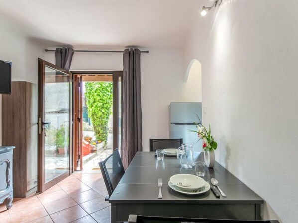 A dining area with a table set for two, a refrigerator, and a view of a garden through an open door. - Le Narcisse by Interhome (Cagnes-sur-Mer)
