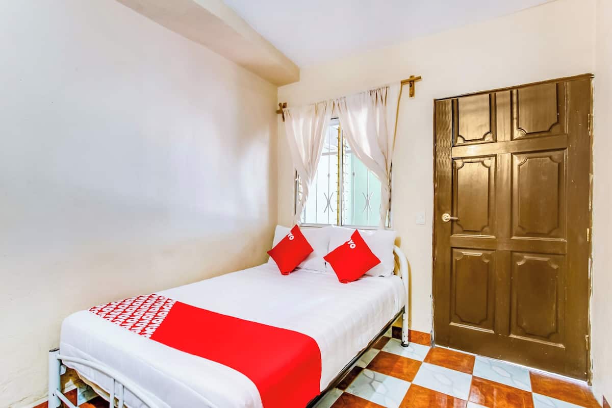 A single bed with a white and red bedspread, two red pillows, and a window with white curtains.