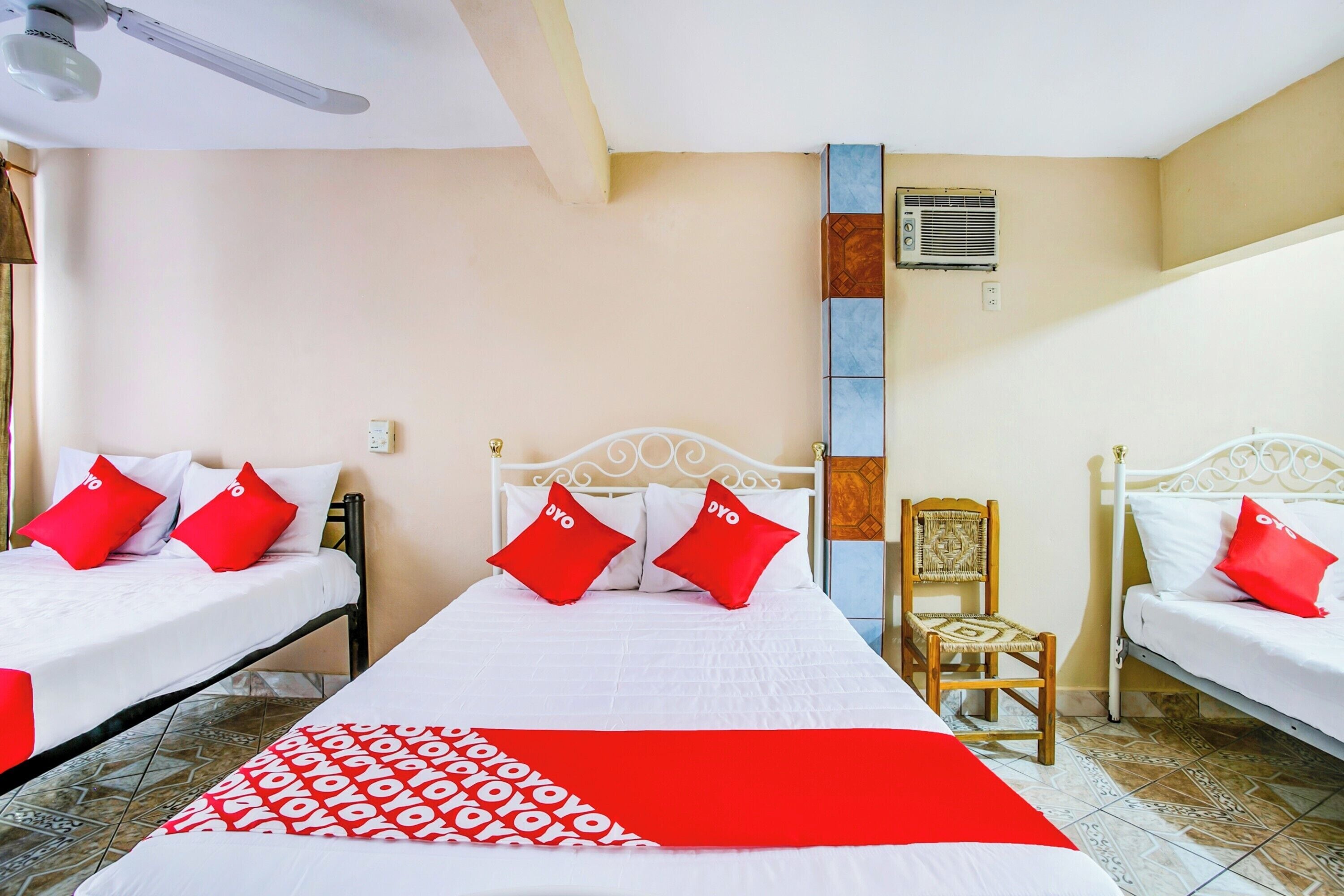 A hotel room with two beds, each with white bedding and red pillows, a red and white patterned throw blanket, a wooden chair, and a wall-mounted air conditioner.