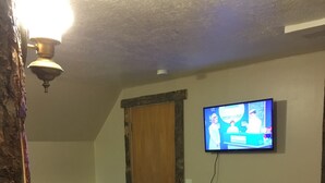 A room with a wooden floor, a television mounted on the wall, a door, and a small seating area with a chair and ottoman. - great for kids - pets welcome - self check in - downtown - kitchen and laundry (Idaho Falls)