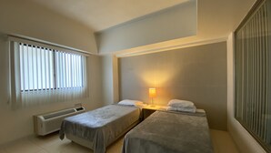 A hotel room with two single beds, a window with blinds, a standing heater, and a nightstand with a lamp. - Pia Resort Hotel Standard 2 Bedroom 1 (Tamuning)