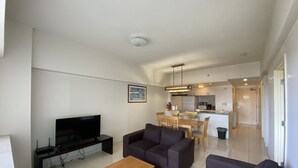 A modern living room with a flat-screen TV, a wooden coffee table, and two sofas. A dining area with a kitchen in the background is visible. - Pia Resort Hotel Standard 2 Bedroom 1 (Tamuning)