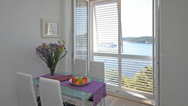 A dining area with a table set for two, a vase of flowers, and a view of the sea through large windows. - Apartment Lavandula - Two-Bedroom Apartment with Sea View (Dubrovnik)