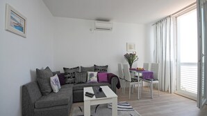 A living room with a grey sofa, a small white table, and a dining area with a glass table and white chairs. - Apartment Lavandula - Two-Bedroom Apartment with Sea View (Dubrovnik)