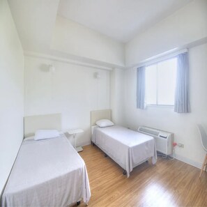 A small, clean room with two single beds, a window with curtains, a radiator, and a chair. - Pia Resort Hotel Corner 2 Bedroom 1 (Tamuning)