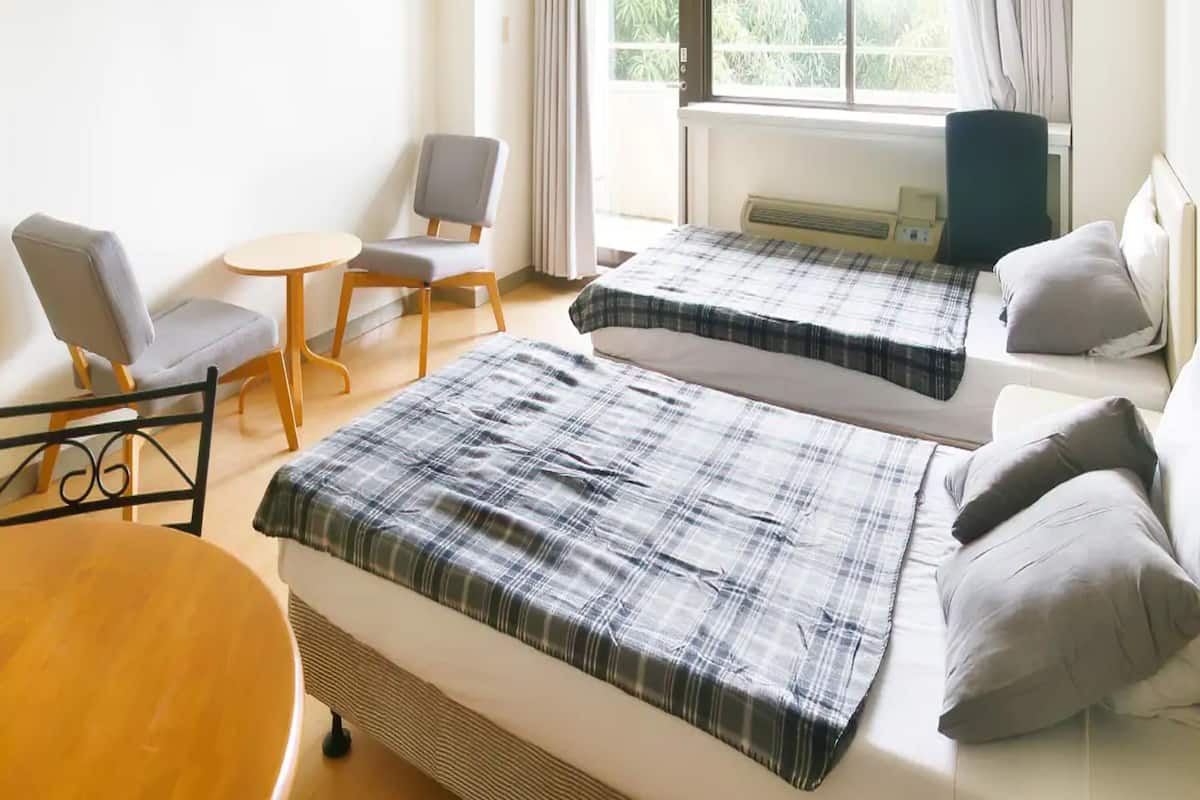 A hotel room with two beds, a small table, a chair, and a window with curtains.