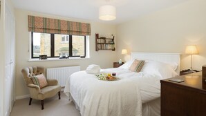 A neatly arranged bedroom with a bed, a chair, a bedside table, a lamp, a bookshelf, and a window with a view of a stone building. - Fairfields, Moreton-in-Marsh, sleeps 6 guests  in 3 bedrooms (Moreton-in-Marsh)