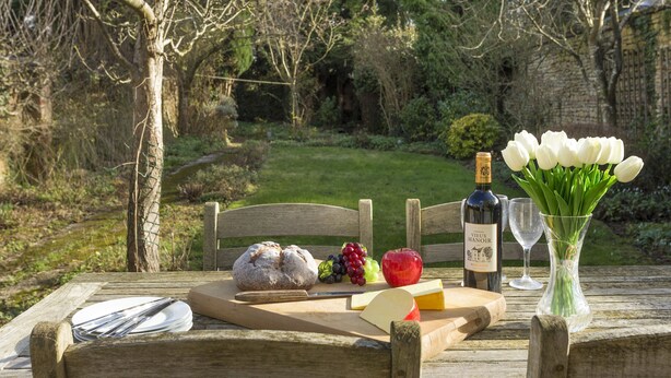 A picnic setup with a loaf of bread, cheese, grapes, an apple, a bottle of wine, and a vase of tulips on a wooden table. - Fairfields, Moreton-in-Marsh, sleeps 6 guests  in 3 bedrooms (Moreton-in-Marsh)