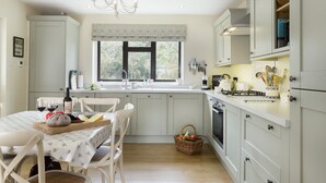 A kitchen with white cabinets, a dining table with chairs, a chandelier, and a window with a view of trees. - Fairfields, Moreton-in-Marsh, sleeps 6 guests  in 3 bedrooms (Moreton-in-Marsh)