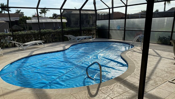 A swimming pool with a blue cover, surrounded by a glass enclosure, and a tiled deck area with lounge chairs. - Gorgeous SW Cape Home on canal w/ pool - Southern Exposure  (Cape Coral)