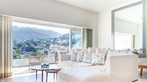 A modern living room with a sofa, a coffee table, and a view of a cityscape and mountains through large windows. - Zenith 601, 2 Balconies, Spectacular Views, in Zona Romantica, Rooftop Pool/Spa (Puerto Vallarta)