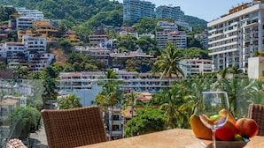 A table with a glass of wine and a bowl of fruit, overlooking a cityscape with buildings and palm trees. - Zenith 601, 2 Balconies, Spectacular Views, in Zona Romantica, Rooftop Pool/Spa (Puerto Vallarta)