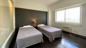 Habitación de hotel con dos camas individuales, un ventanal con persianas, cabecera oscura y unidad de aire acondicionado visible.