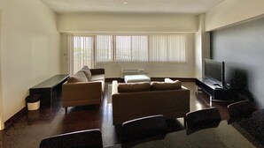 Una sala de estar con un sofá, un televisor de pantalla plana y un amplio ventanal con persianas.
