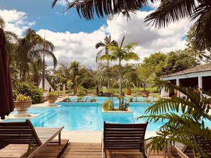 A pool area with wooden deck chairs, palm trees, and a clear blue sky. - Naina Park Resort (La Foa)