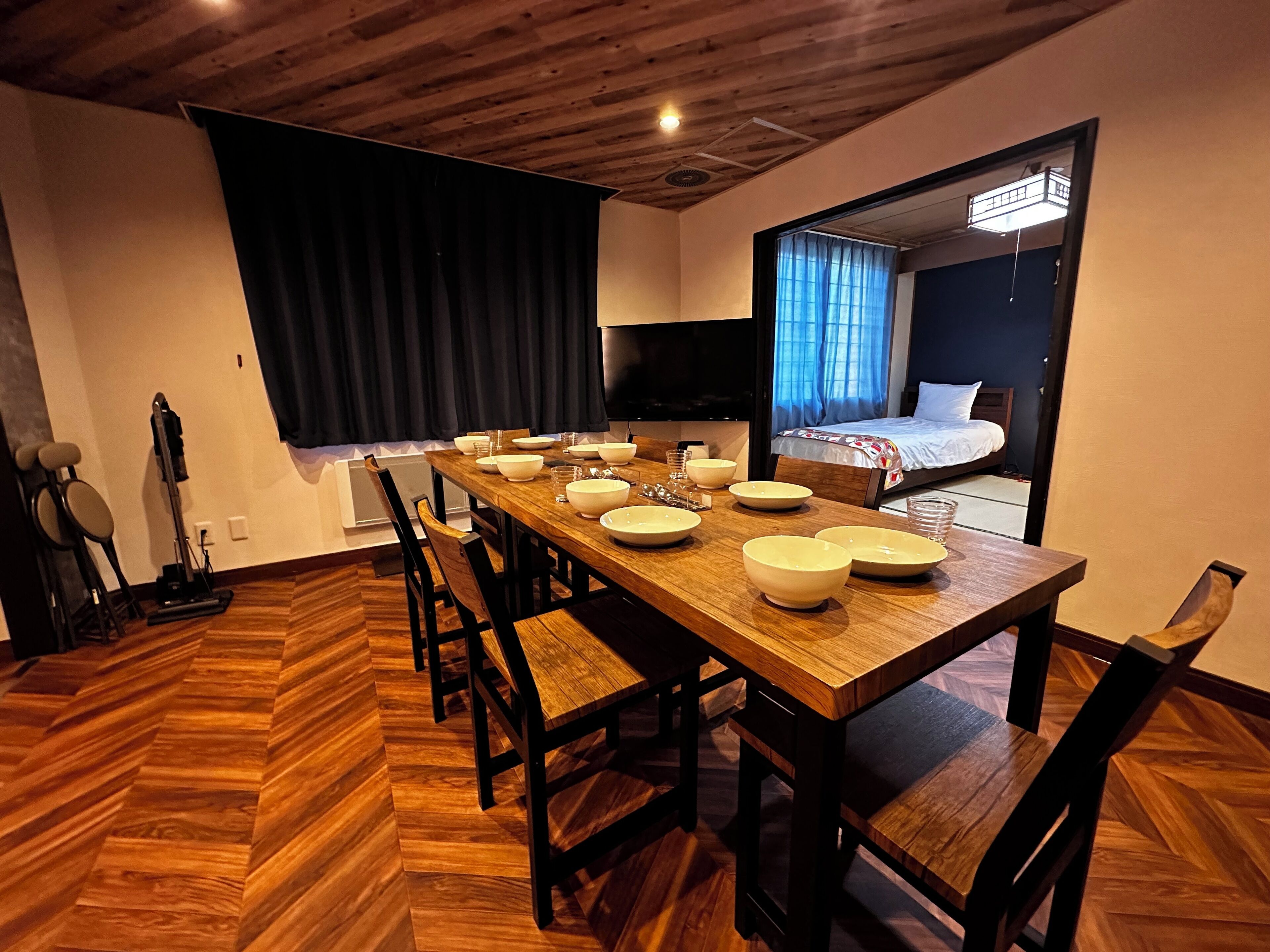 Une chambre d’hôtel moderne avec une table à manger en bois, dressée pour quatre personnes, un grand lit et un téléviseur fixé au mur.