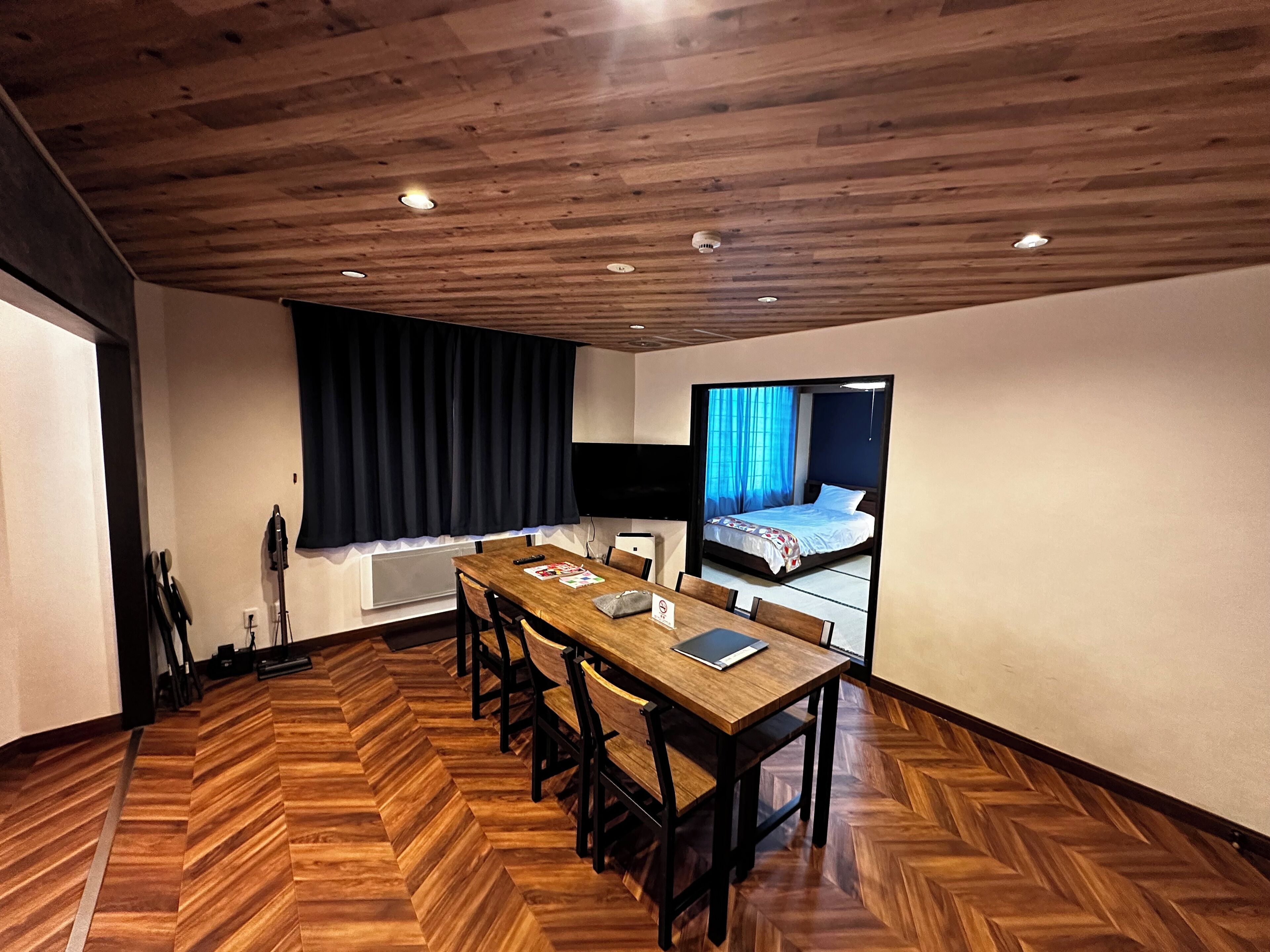 Ein Zimmer mit Holzdielen im Fischgrätmuster, einem Holzesstisch mit Stühlen, einem Bett mit blauer Vorhang und einem an der Wand befestigten Flachbildfernseher.