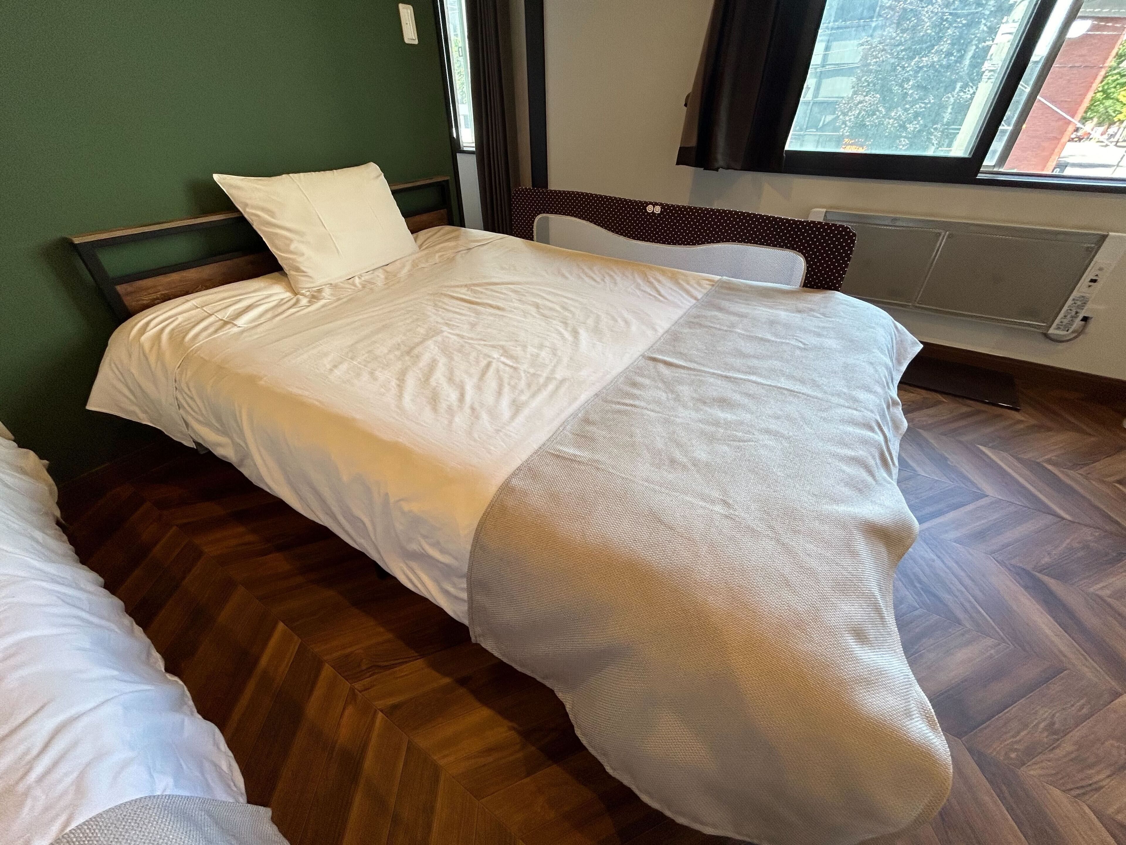 A bed with a white comforter and pillows, a wooden floor, a green wall, and a window with curtains.
