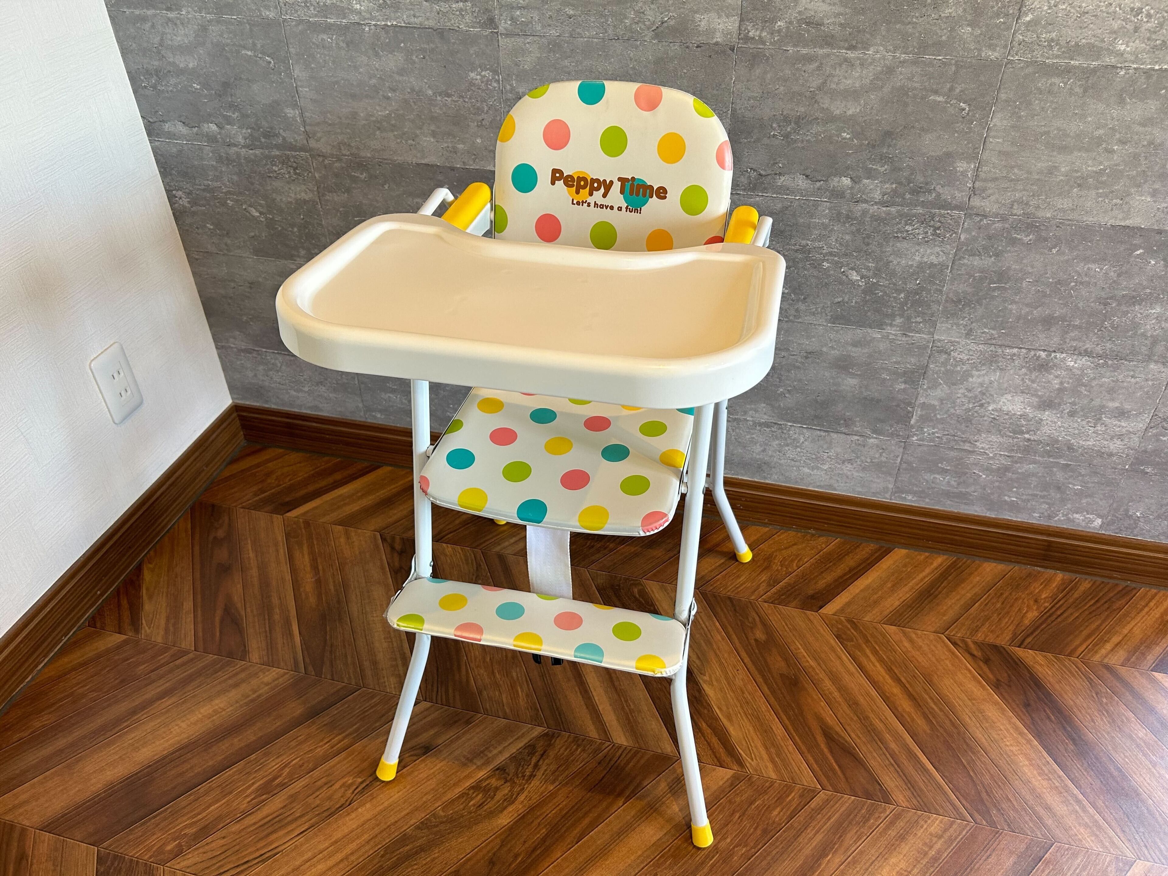A baby high chair with a polka dot design and a tray, placed on a wooden floor with a herringbone pattern.
